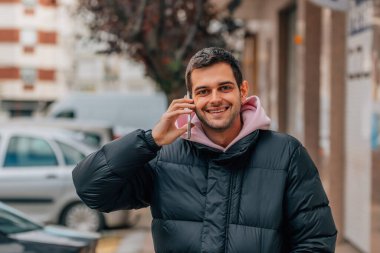 Sokaktaki genç adam kışlık giysiler içinde cep telefonuyla konuşuyor.