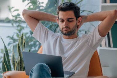 Genç adam evinde bilgisayar başında kanepede dinleniyor.