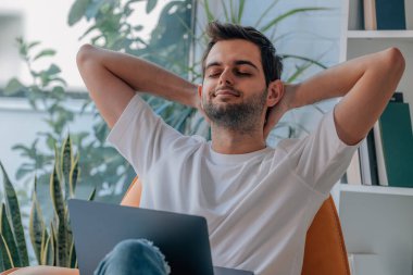 Genç adam evinde bilgisayar başında kanepede dinleniyor.