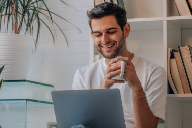 Evinde bilgisayar ve kahveyle oturan genç adam.