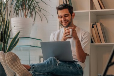 Evinde bilgisayar ve kahveyle oturan genç adam.