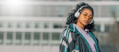 Kulaklıklı ve güneş gözlüklü kız sokakta müzik dinliyor.