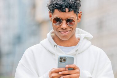 Güneş gözlüklü genç adam sokakta cep telefonuna bakıyor.