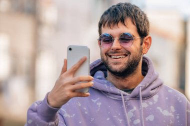 Genç adam güneş gözlüklü cep telefonuna bakıyor.