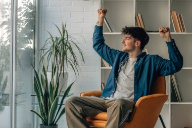 young male on the sofa at home with arms up celebrating