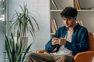 Evdeki kanepede oturan genç adam cep telefonuyla.