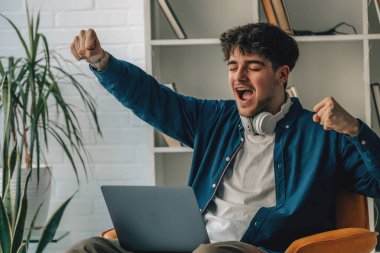 Evde dizüstü bilgisayarı olan genç bir adam kanepeye oturmuş neşe ve başarıyla heyecanlanıyor.