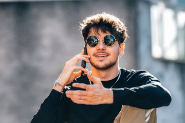 Dışarıda güneş gözlüklü ve cep telefonlu genç bir adam var.
