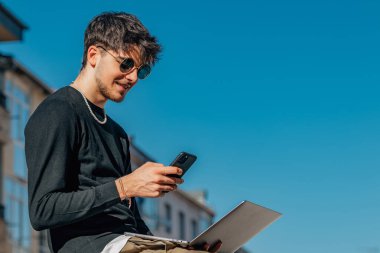 Sokakta dizüstü bilgisayarı ve cep telefonu olan genç bir adam.