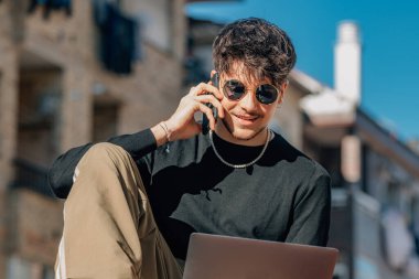 Açık terasta cep telefonu ve dizüstü bilgisayarı olan genç bir adam.