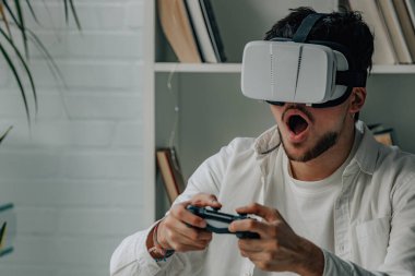 Evde elinde joystick ve sanal gerçeklik gözlükleriyle oyun oynayan genç bir adam.