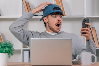 Evde dizüstü bilgisayarı olan genç bir erkek cep telefonuna hayret içinde bakıyor.