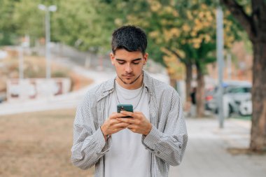 Genç bir erkek sokakta yürürken cep telefonuna ya da akıllı telefonuna bakıyor.