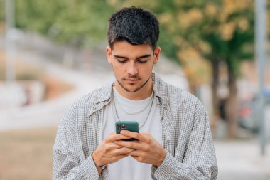 Genç bir erkek sokakta yürürken cep telefonuna ya da akıllı telefonuna bakıyor.
