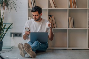 Evdeki genç adam dizüstü bilgisayarlı telefon ve bir fincan kahveyle rahatladı.