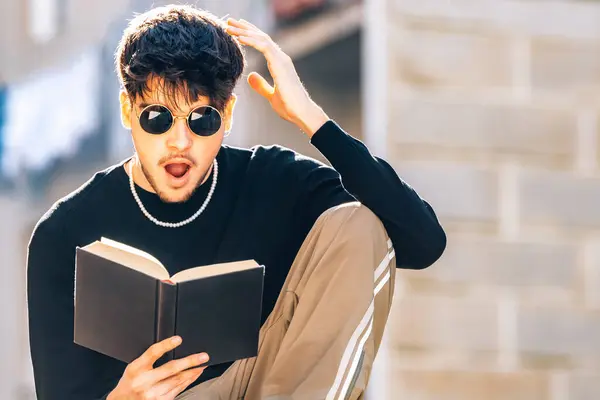 young man reading with textbook outdoors with surprised expression