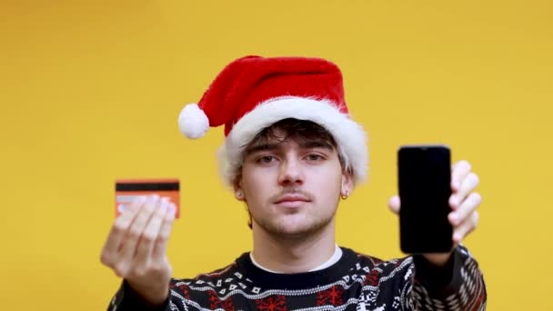 サンタクロースの帽子をかぶったクレジットカードと携帯電話を持つ若者 クリスマスショッピング — ストック動画