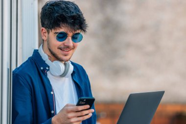 Dışarıda güneş gözlüğü takan cep telefonu ve dizüstü bilgisayarı olan genç bir adam.