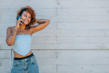 Latin Afrikalı Amerikalı dövmeli bir kız cep telefonuyla konuşuyor, sokak duvarında izole edilmiş.