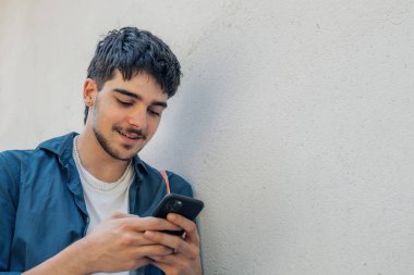 Duvara yaslanmış cep telefonuna bakan genç adam.