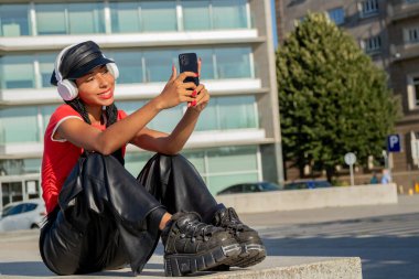 Kulaklık ve cep telefonuyla sokakta oturan bir kız.