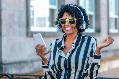 Kulaklıklı ve cep telefonlu bir kadın sarı gözlüklü gülümsüyor.