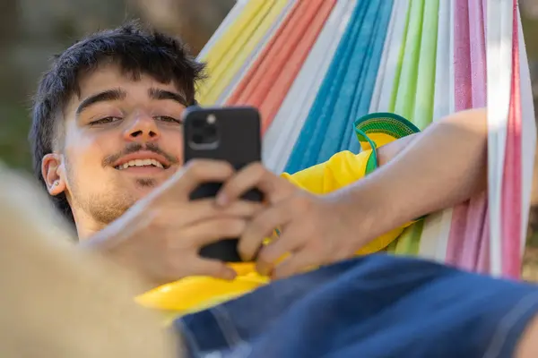 夏天坐在吊床上的年轻人用手机放松了下来 免版税图库照片