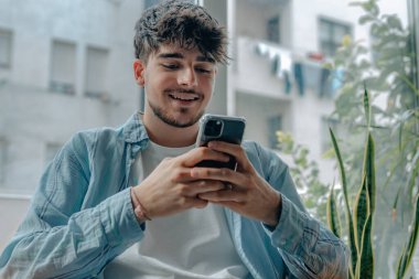 Cep telefonlu genç adam evdeki kanepede oturuyor.