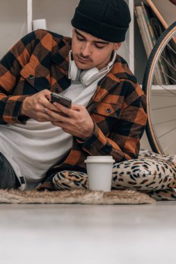 Cep telefonlu, şapkalı ve sonbahar kış kıyafetleriyle evde oturan genç adam.