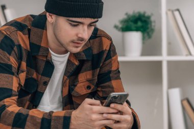 Evde oturan genç adam cep telefonuna bakıyor.