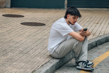 Genç adam sokakta oturuyor ve cep telefonuna bakıyor.