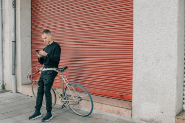 Sokakta kırmızı arka planda cep telefonu ve bisikleti olan genç adam.