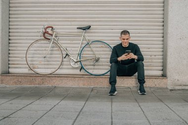 Genç adam cep telefonuyla sokakta oturuyor.