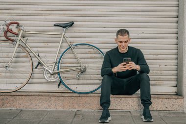 Genç adam cep telefonuyla sokakta oturuyor.