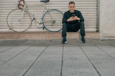 Genç adam cep telefonuyla sokakta oturuyor.