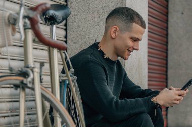 Genç adam cep telefonuyla sokakta oturuyor.