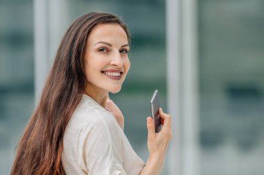 İş kadını cep telefonuyla kameraya bakıp gülümsüyor.