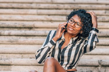 Latin Afrikalı Amerikalı kadın sokakta cep telefonuyla oturuyor.