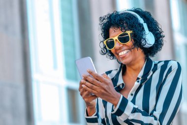 Kulaklıklı ve cep telefonlu bir kadın sarı gözlüklü gülümsüyor.