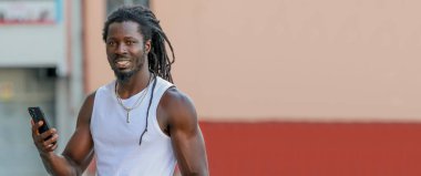 Afro saçlı genç bir adam. Sokakta cep telefonuna bakıyor.