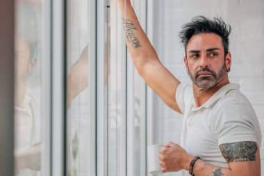 Sakallı çekici bir adam pencerede kahve içiyor.