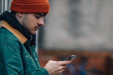 Şapkalı ve kalın elbiseli genç bir adamın portresi cep telefonuna bakıyor.