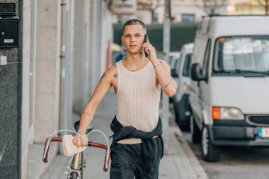 Elinde bisikletle sokakta yürüyen genç bir adam cep telefonuyla konuşuyor.