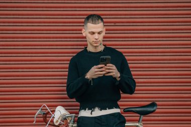 Sokakta kırmızı arka planda cep telefonu ve bisikleti olan genç adam.