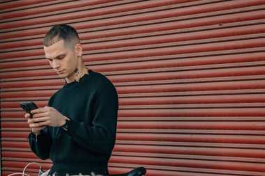 Sokakta kırmızı arka planda cep telefonu ve bisikleti olan genç adam.