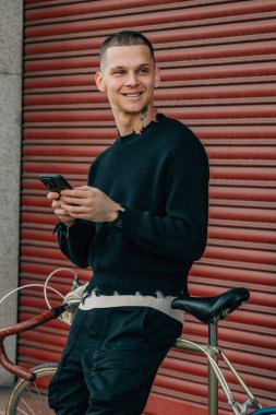 Sokakta kırmızı arka planda cep telefonu ve bisikleti olan genç adam.