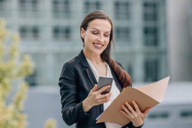 Sokakta doküman klasörlü iş kadını cep telefonuna bakıyor