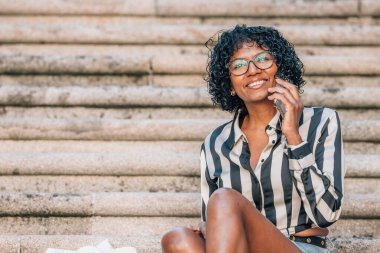 Latin Afrikalı Amerikalı kadın sokakta cep telefonuyla oturuyor.