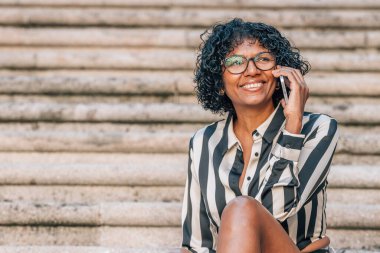 Latin Afrikalı Amerikalı kadın sokakta cep telefonuyla oturuyor.