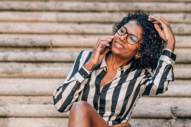 Latin Afrikalı Amerikalı kadın sokakta cep telefonuyla oturuyor.